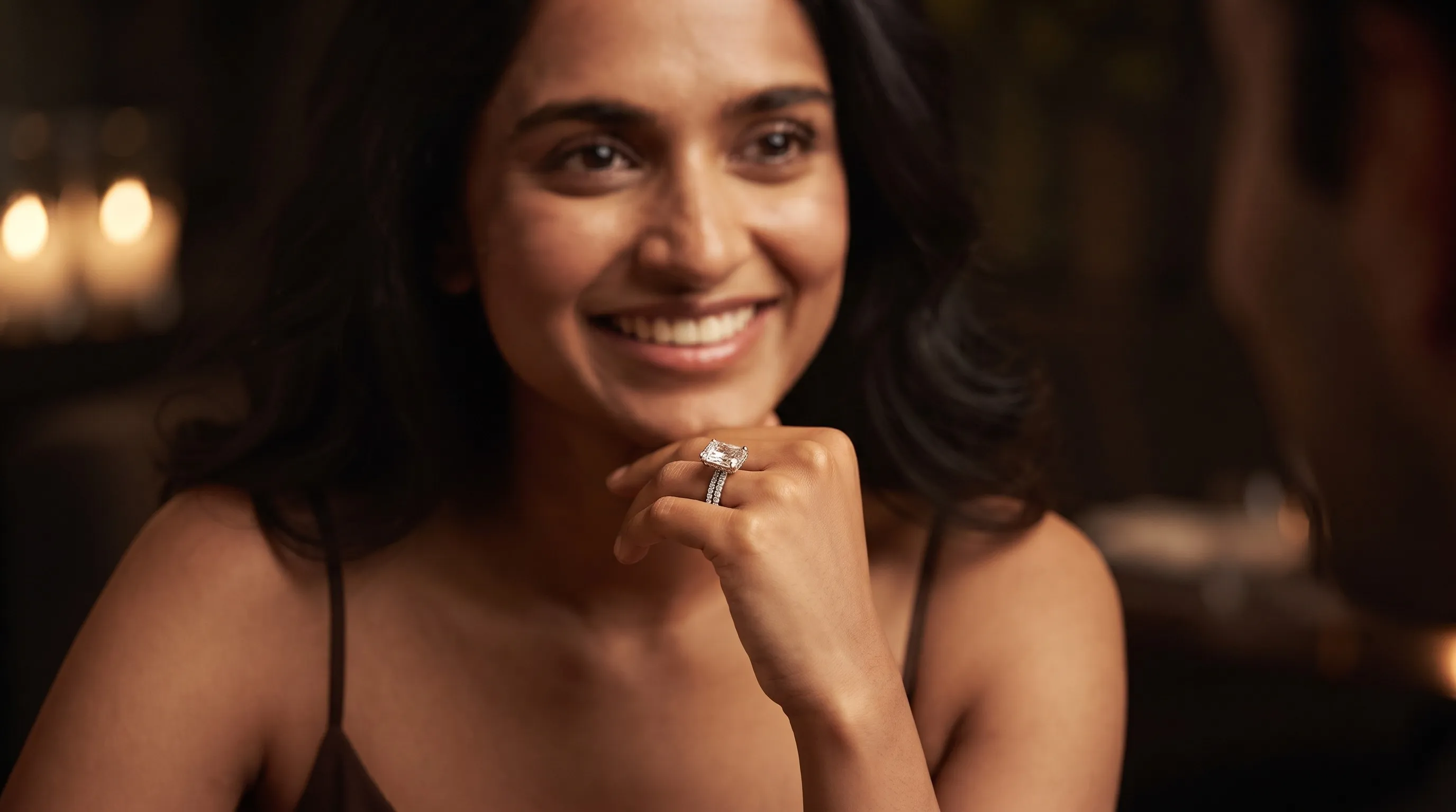 A woman smiling with her chin on her hand, wearing an emerald-cut solitaire ring on a pave band, in a candlelit room.