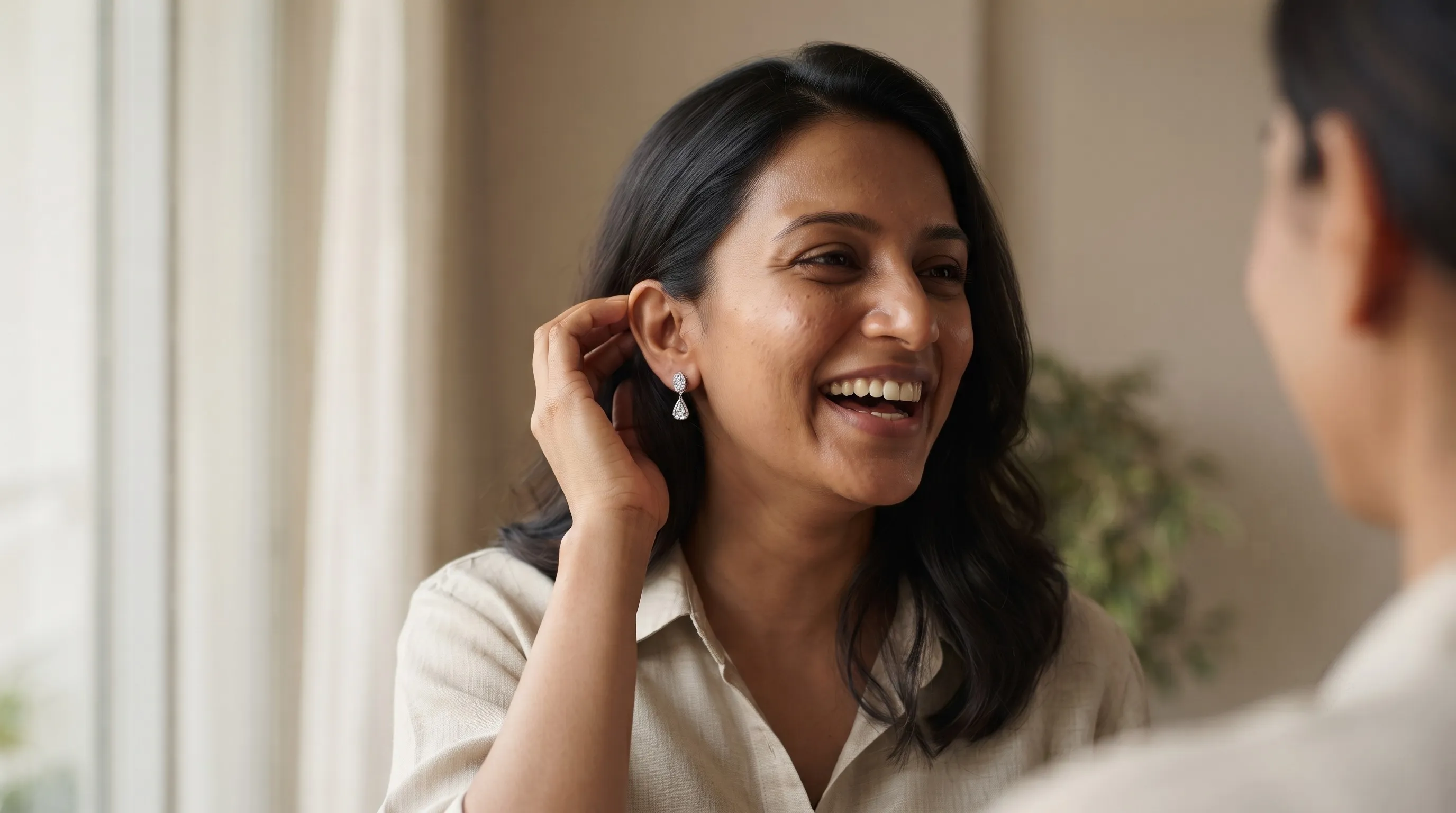 A woman laughing as she tucks her hair behind her ear, wearing a pear-shaped drop earring, in a bright room.