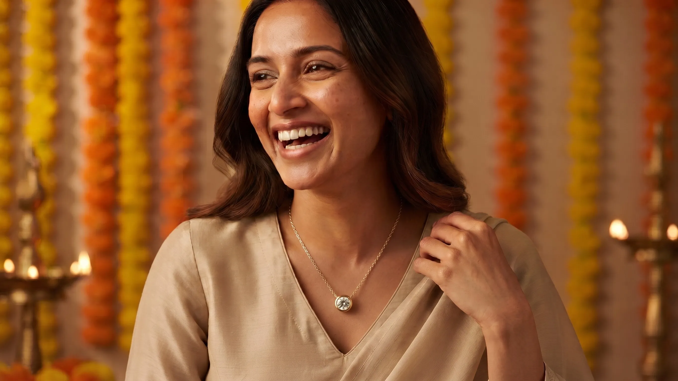 A woman in a beige silk kurta laughing, wearing a bezel-set round pendant, with marigold garlands and oil lamps behind her.